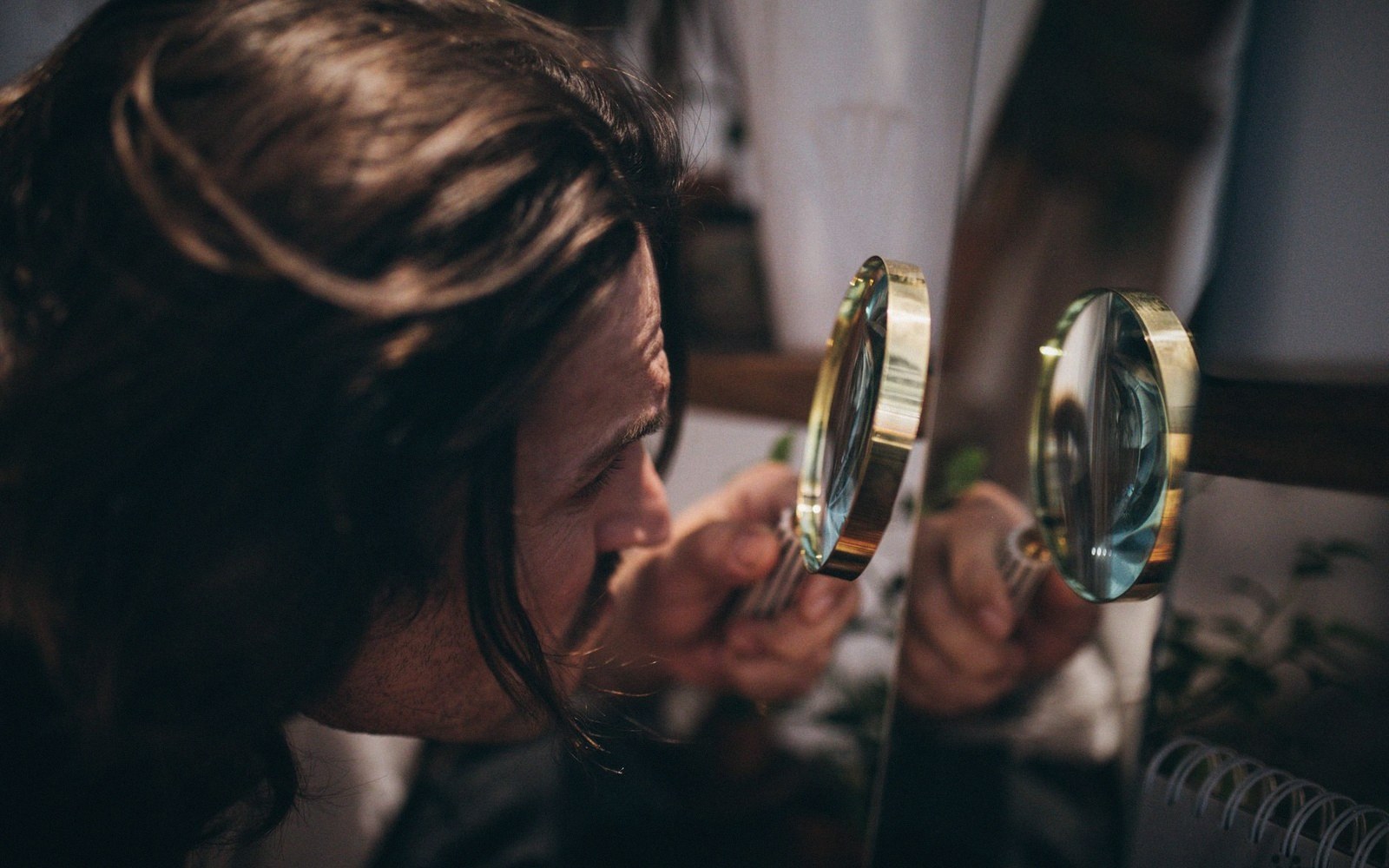 A man with long hair examines an object closely through a brass magnifying glass