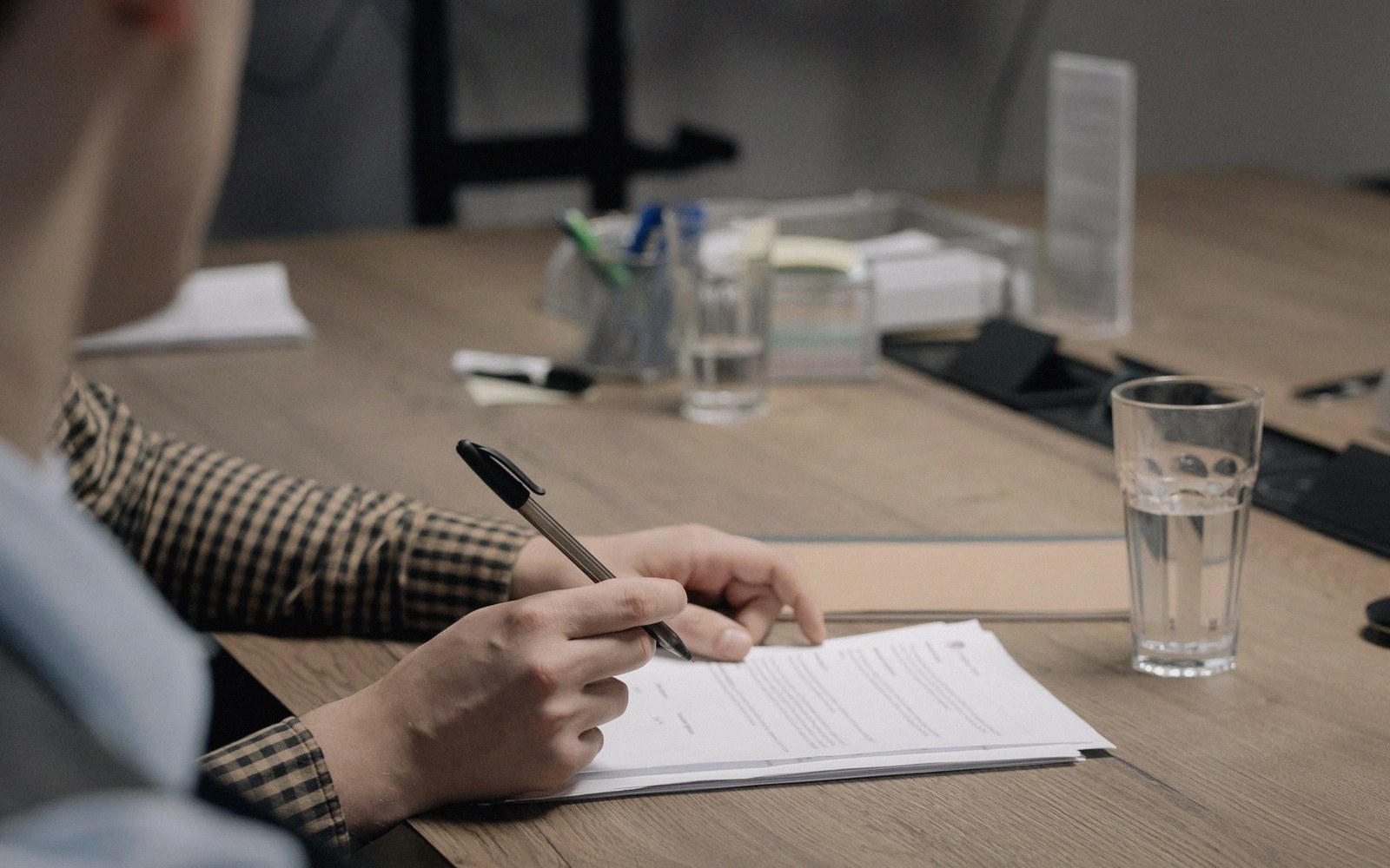 A person signs a document at a wooden desk with a glass of water nearby