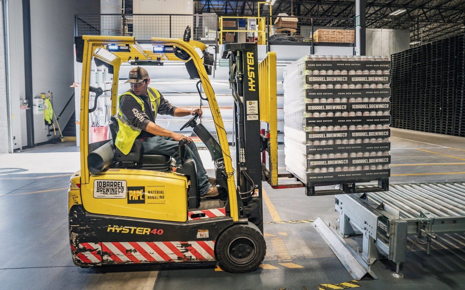 Worker drives a forklift moving pallets of canned beer in a brewery warehouse
