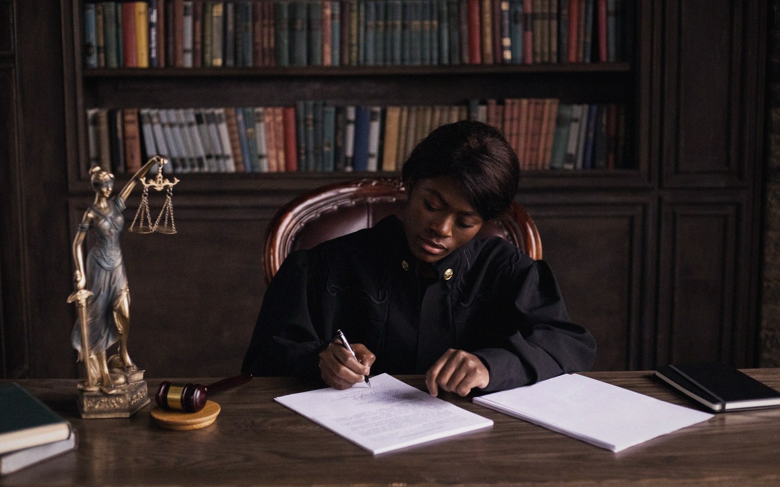 A judge in robes signs documents at a desk beside a gavel and justice statuette