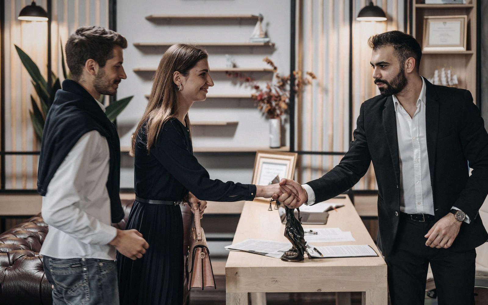 A woman shakes hands with a suited man across a desk as another man watches