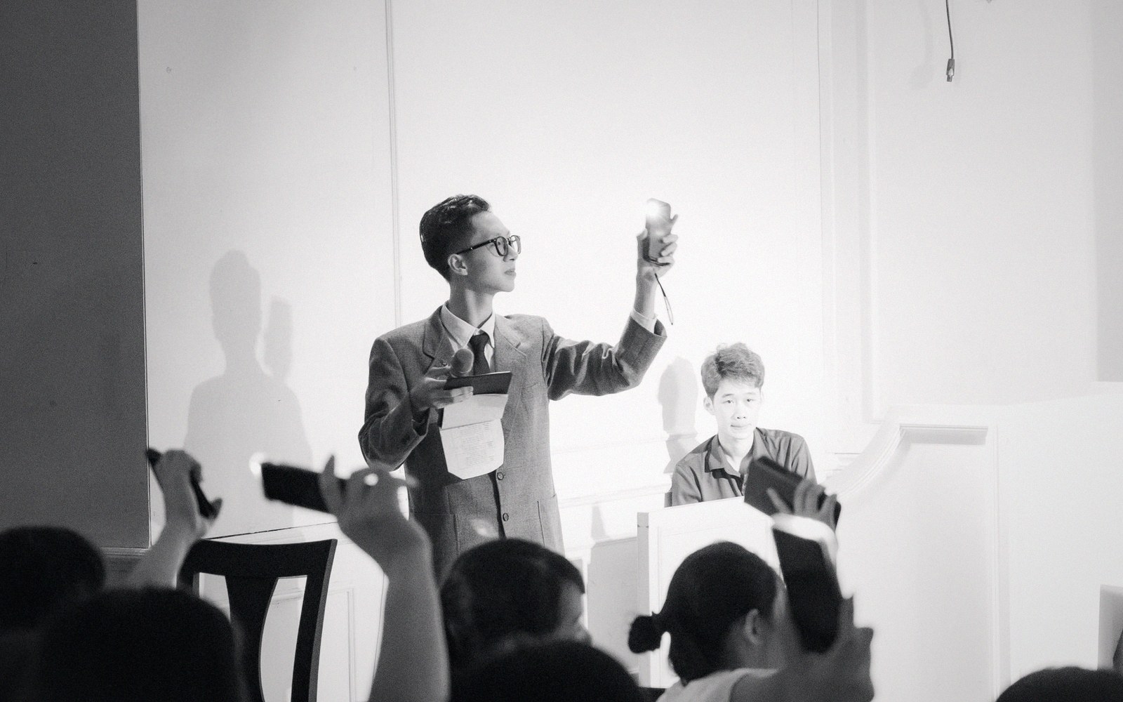 Man in glasses raises a small object during a presentation as onlookers film with phones