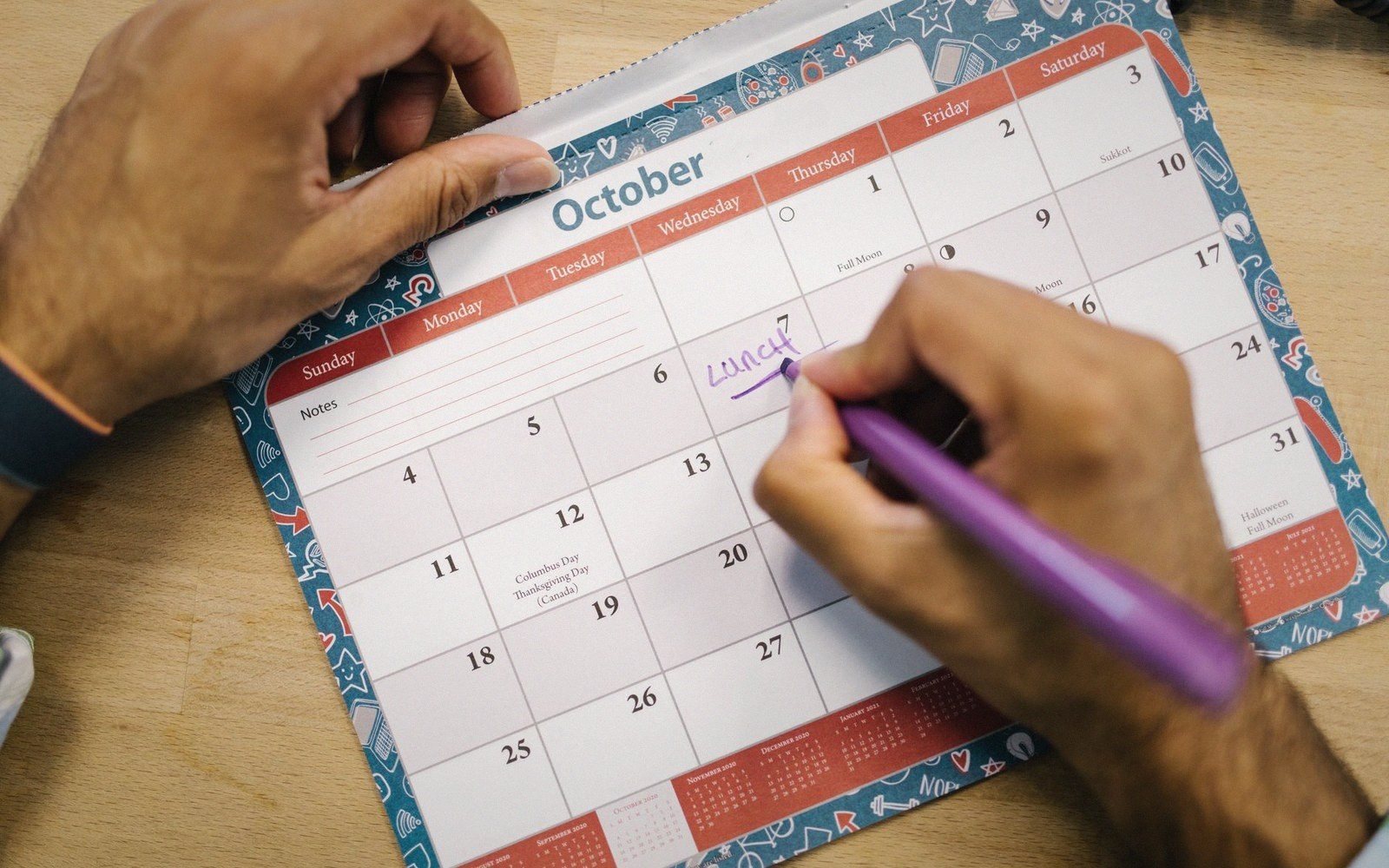 A hand writes "lunch" on a paper October calendar with a purple pen
